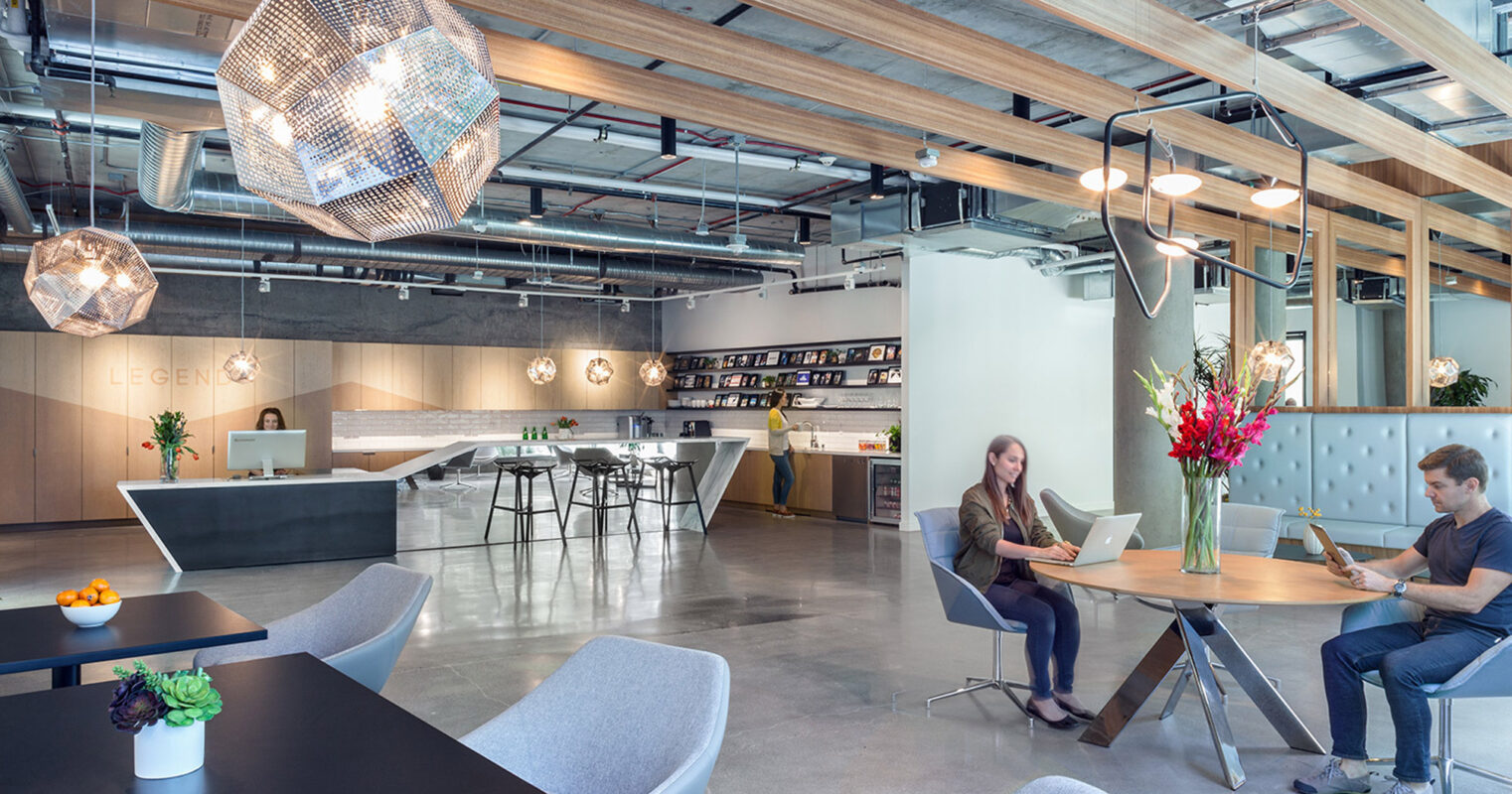 Modern office space showcasing exposed ceiling beams and ductwork, with a large geometric pendant light. Features an open floor plan with a kitchenette, communal dining area, and casual seating. A mix of natural and artificial lighting enhances the industrial yet inviting ambiance.