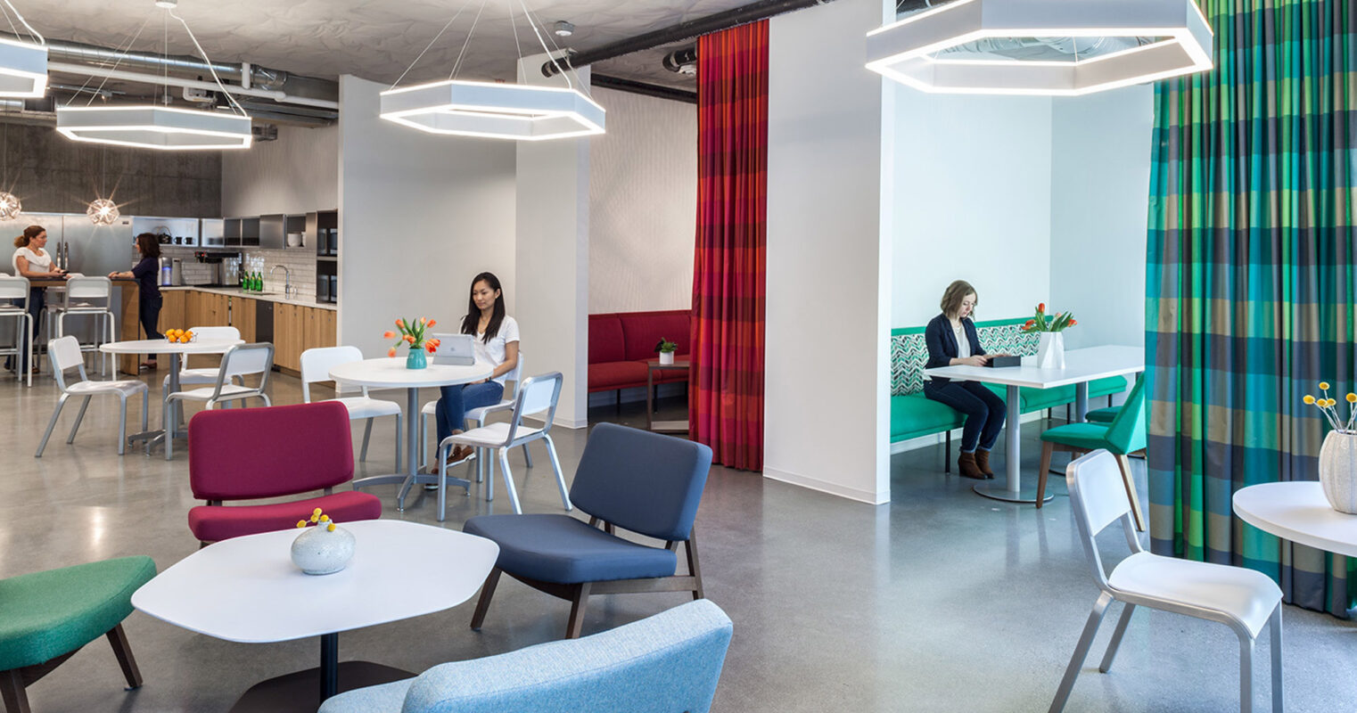 Modern office break area with varied seating options including booths and chairs, accentuated by vibrant color contrasts, geometric light fixtures, and open-plan greenery for a contemporary, inviting space.