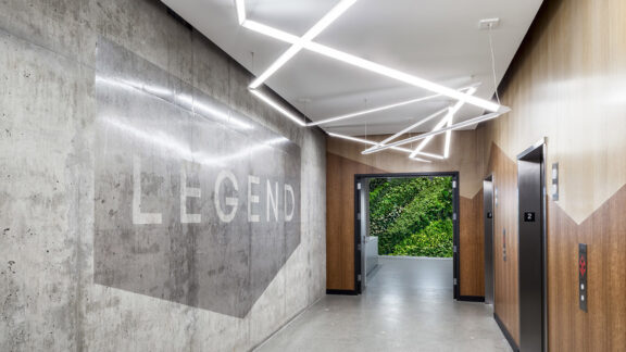 Modern hallway with geometric LED ceiling lights and polished concrete walls featuring a bold stenciled "LEGEND". Warm wooden doors contrast with the industrial aesthetics, complemented by a lush living green wall at the end of the corridor.