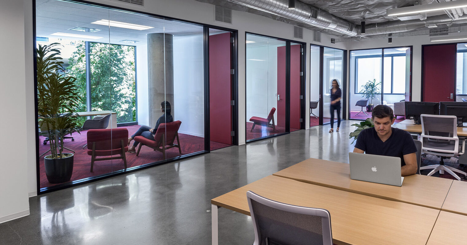 Modern office interior with floor-to-ceiling glass partitions, framing a breakout space with red armchairs and a coffee table. Strategically placed greenery complements the natural light, while a focused individual works at a sleek, communal wooden table in the foreground.