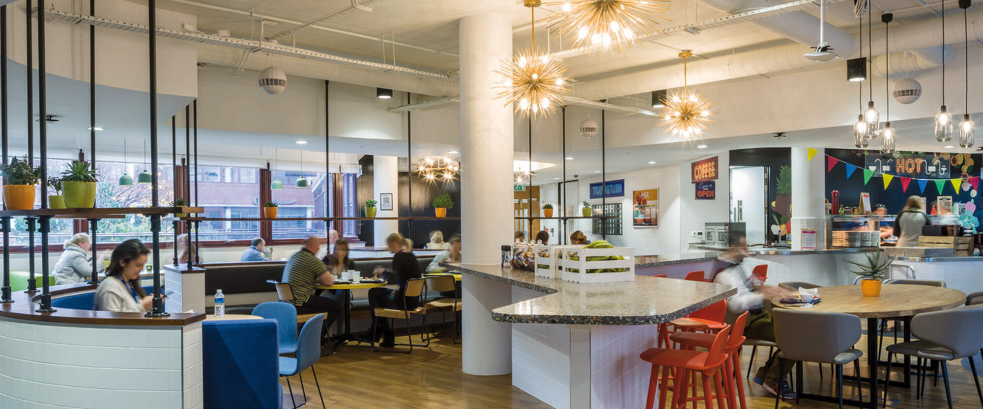 Spacious café interior with contemporary style, featuring an eclectic mix of seating arrangements, vibrant pendant lighting, and a white curved service counter. Exposed ductwork and concrete ceiling provide an industrial contrast to the warm wood tones and colorful decor.