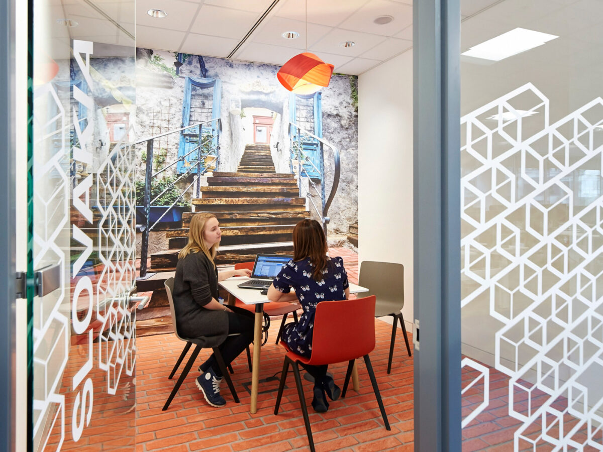 Two individuals engage in discussion at a modern workspace, featuring eclectic chairs and a large mural of a rustic staircase, complemented by geometric translucent window decals.