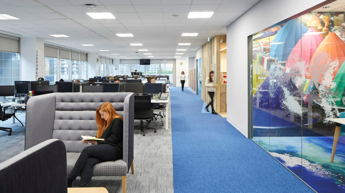 Modern open-plan office space with ergonomic chairs and workstations, accented by vibrant blue carpeting. High-backed gray acoustic sofas provide informal meeting spots, while colorful abstract wall art contributes to an energetic atmosphere. Glass-walled meeting rooms enhance the sense of openness and connectivity.