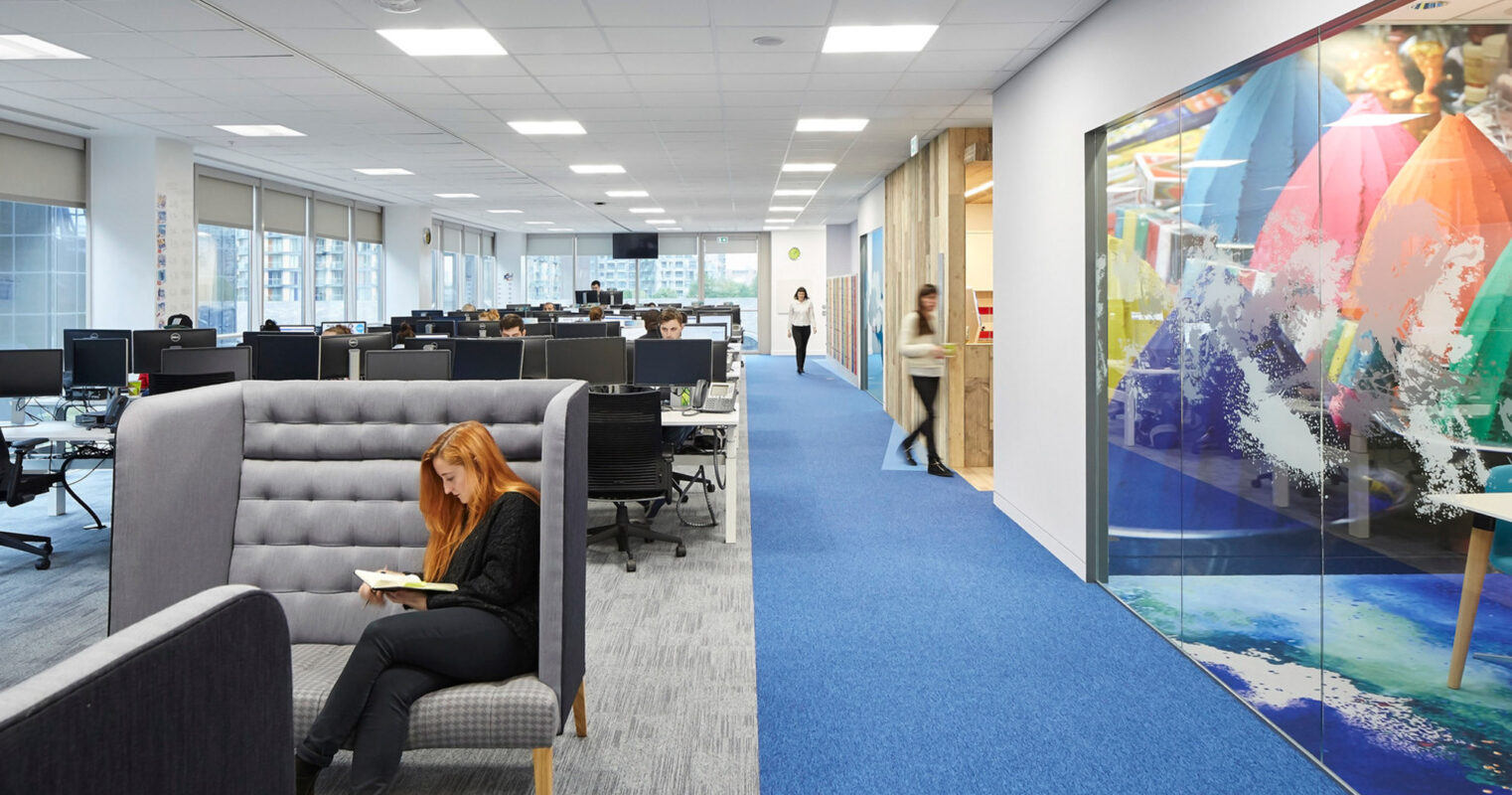 Modern open-plan office space with ergonomic chairs and workstations, accented by vibrant blue carpeting. High-backed gray acoustic sofas provide informal meeting spots, while colorful abstract wall art contributes to an energetic atmosphere. Glass-walled meeting rooms enhance the sense of openness and connectivity.