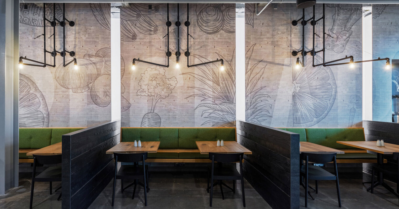 Modern industrial-style dining space featuring exposed ceiling pipes, concrete walls adorned with botanical illustrations, and an eclectic mix of hanging pendant lights. Dark wooden tables complement green upholstered booth seating, creating a harmonious, chic urban eatery ambiance.