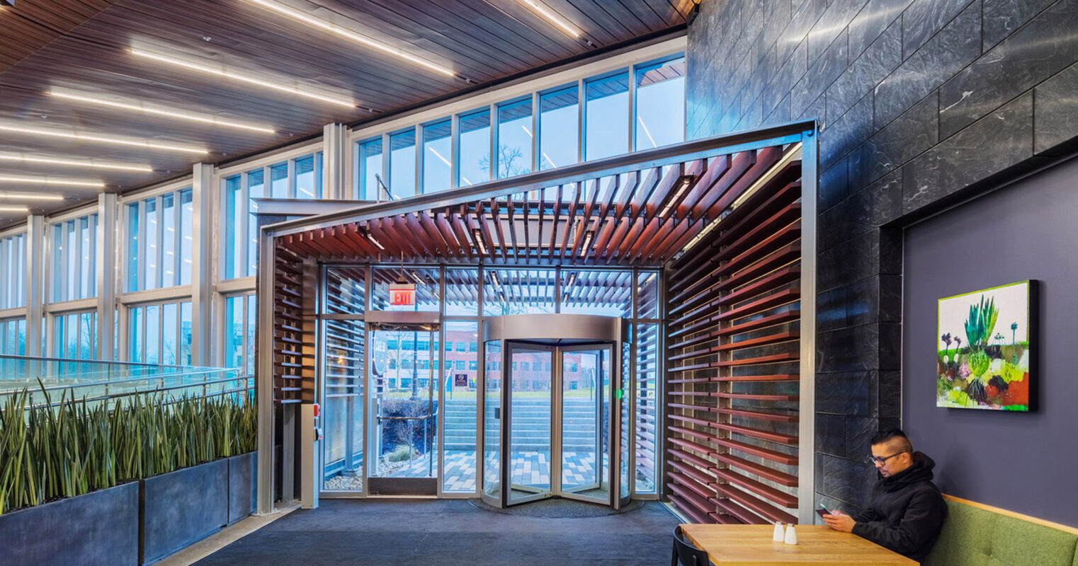 Modern commercial lobby featuring sleek wooden slats on the ceiling that lead to a luminous, glass-enclosed entrance, complementing the textured dark stone wall. Green upholstered bench seating adds a pop of color beside a vibrant wall art piece, while a patron works at a table nearby.