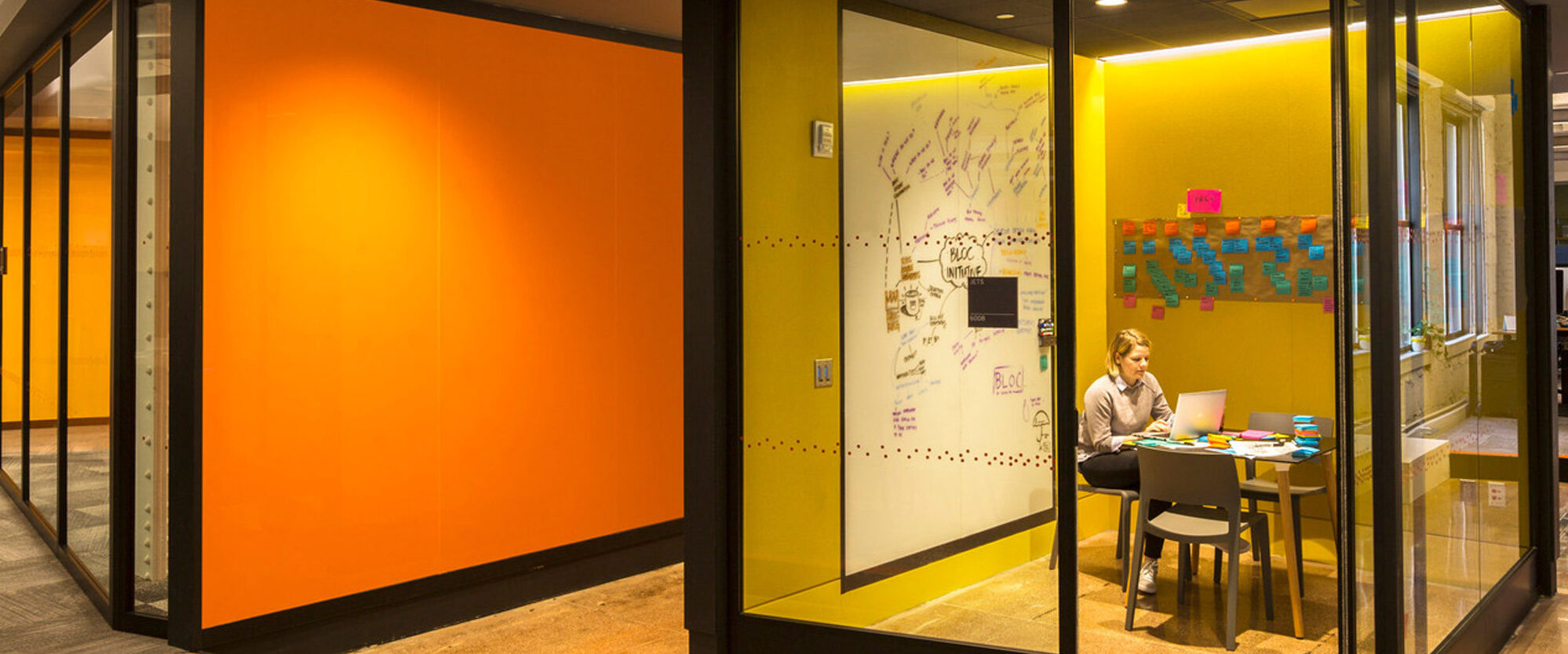 Modern office space with vibrant orange and yellow hues featuring glass-partitioned meeting rooms. A person works at a desk with post-it notes on the wall, suggesting creative brainstorming within a contemporary design.