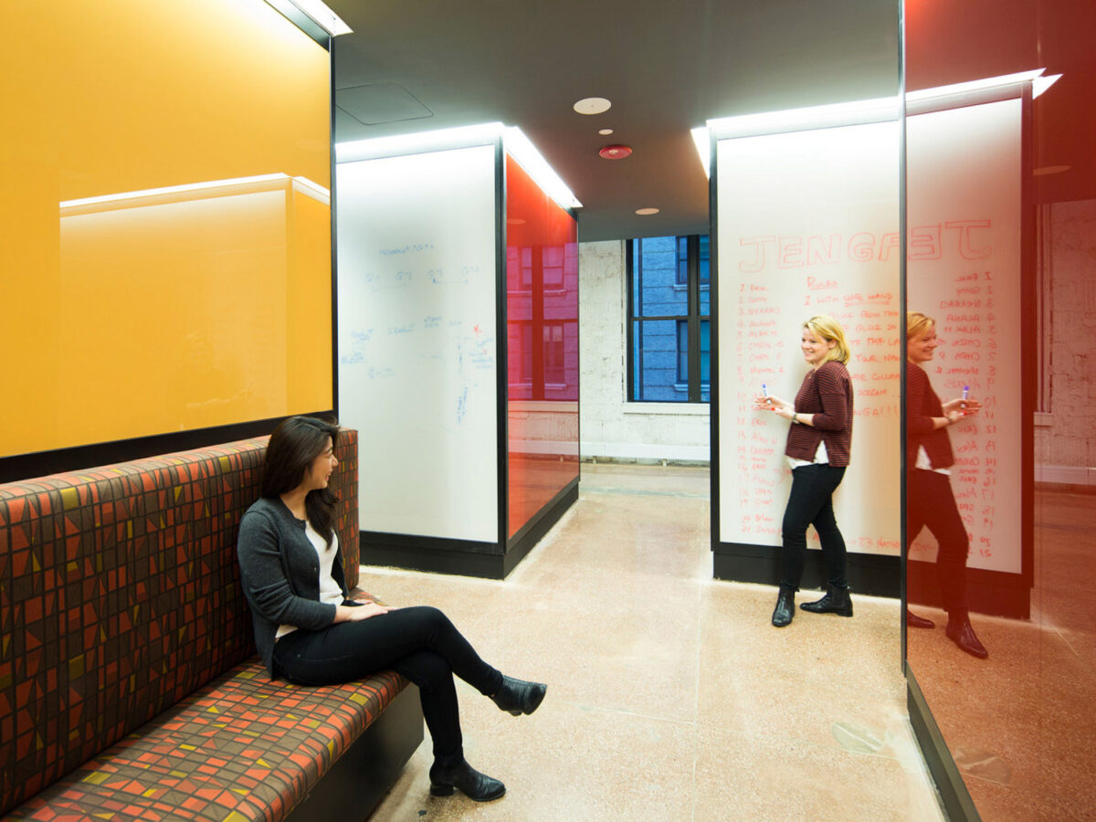Contemporary office lounge with vibrant orange walls, plaid upholstered bench seating, and whiteboard-coated glass partitions used for collaborative brainstorming. Natural light filters through the large window, enhancing the lively ambiance.