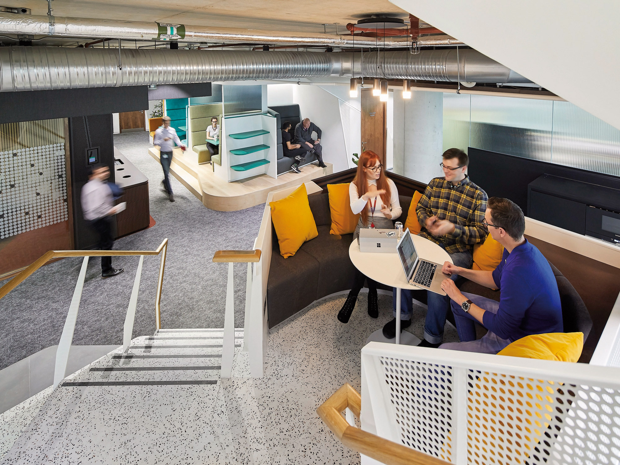 Modern office space with industrial aesthetic, featuring exposed ductwork and a speckled terrazzo floor. A central wooden table with yellow accents provides a collaborative work area, while a slide in the background adds playful functionality. Mesh room dividers offer privacy without visual obstruction.