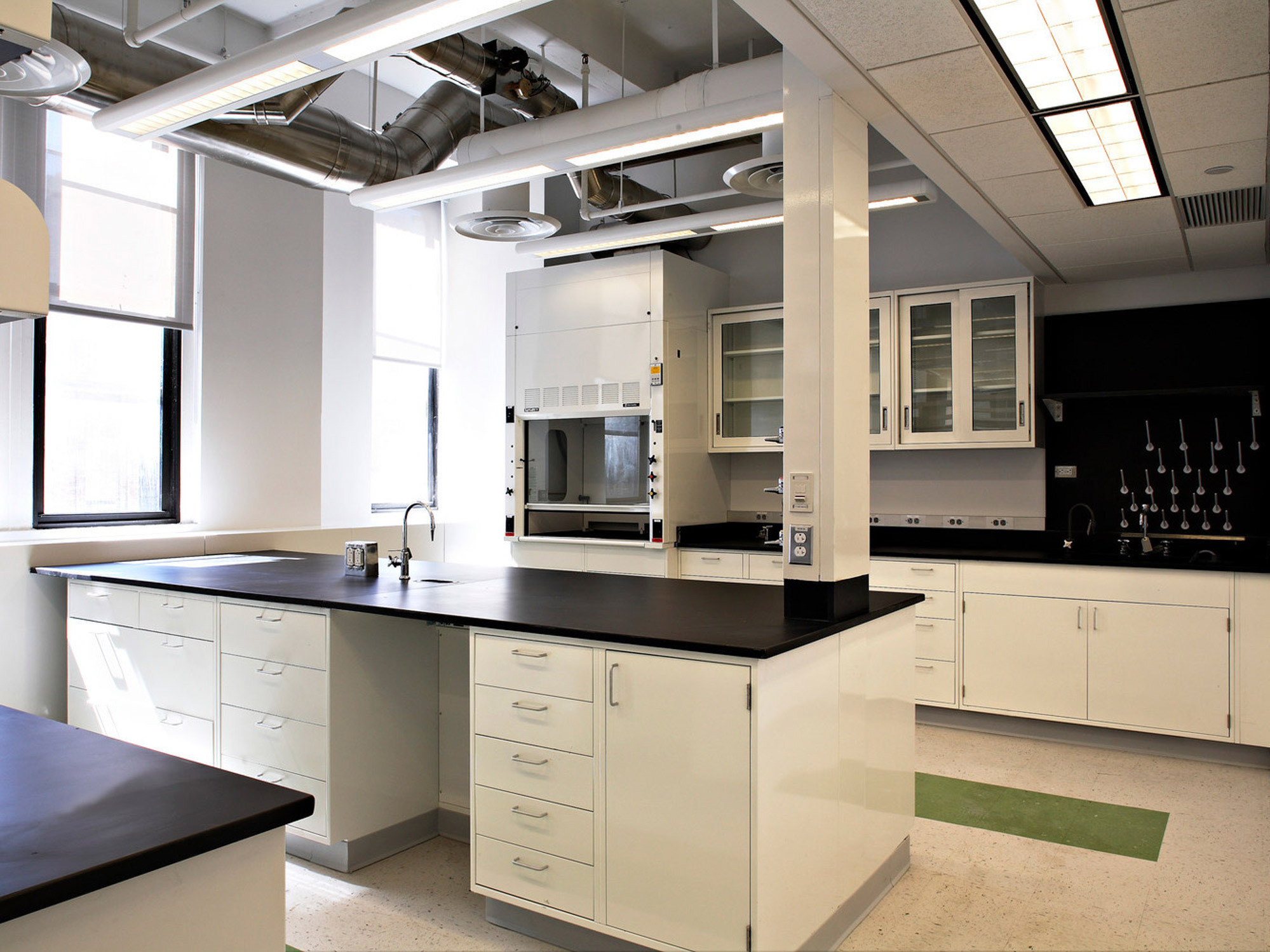 Modern laboratory interior with clean lines, featuring white cabinetry and a sleek black countertop. Overhead, exposed ductwork adds an industrial touch, while track lighting ensures a well-lit workspace. Green floor markings guide safety paths.