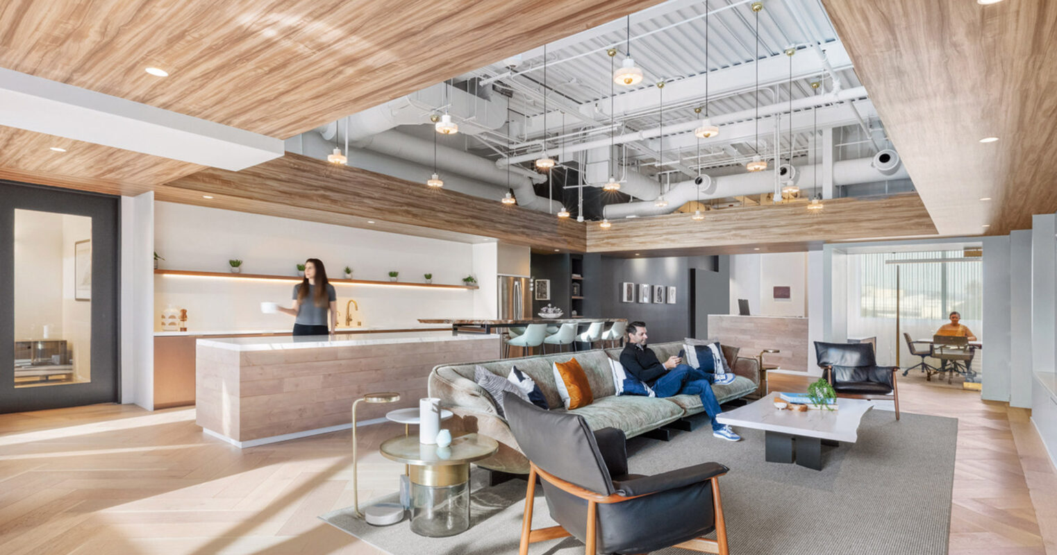 Modern open-plan office lounge with herringbone wood ceiling, polished concrete floor, and central coffee bar. Black leather armchairs, glass-top tables, and subtle beige carpet define the waiting area, with abundant natural light from large windows enhancing the warm, inviting atmosphere.