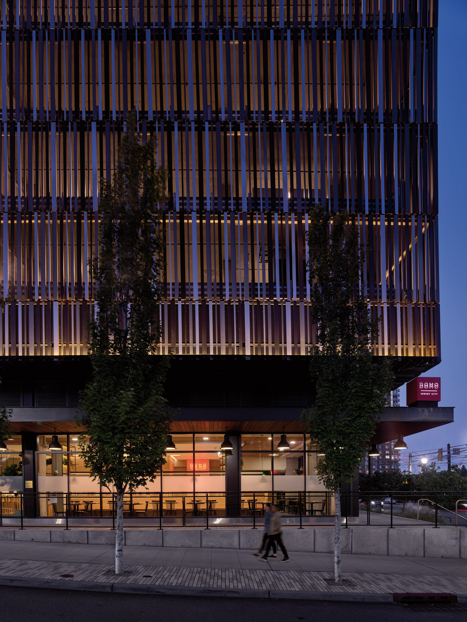 Modern architectural building at dusk showcasing vertical timber louvers for sun shading, with the ground floor featuring transparent glass windows offering a glimpse into the illuminated interior space. A pedestrian walks by, emphasizing the blend of urban environment and innovative design.
