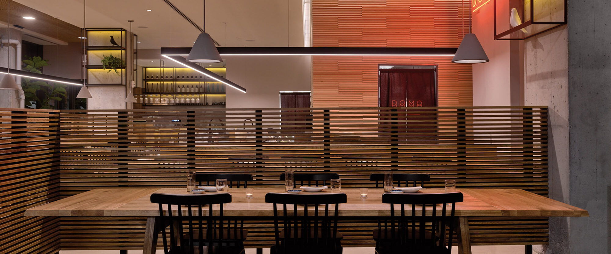 Modern dining space featuring a wooden communal table with black chairs, pendant lighting, and slatted wood partitions. Warm-toned backlighting accentuates the textured rear wall, creating a cozy, inviting atmosphere.