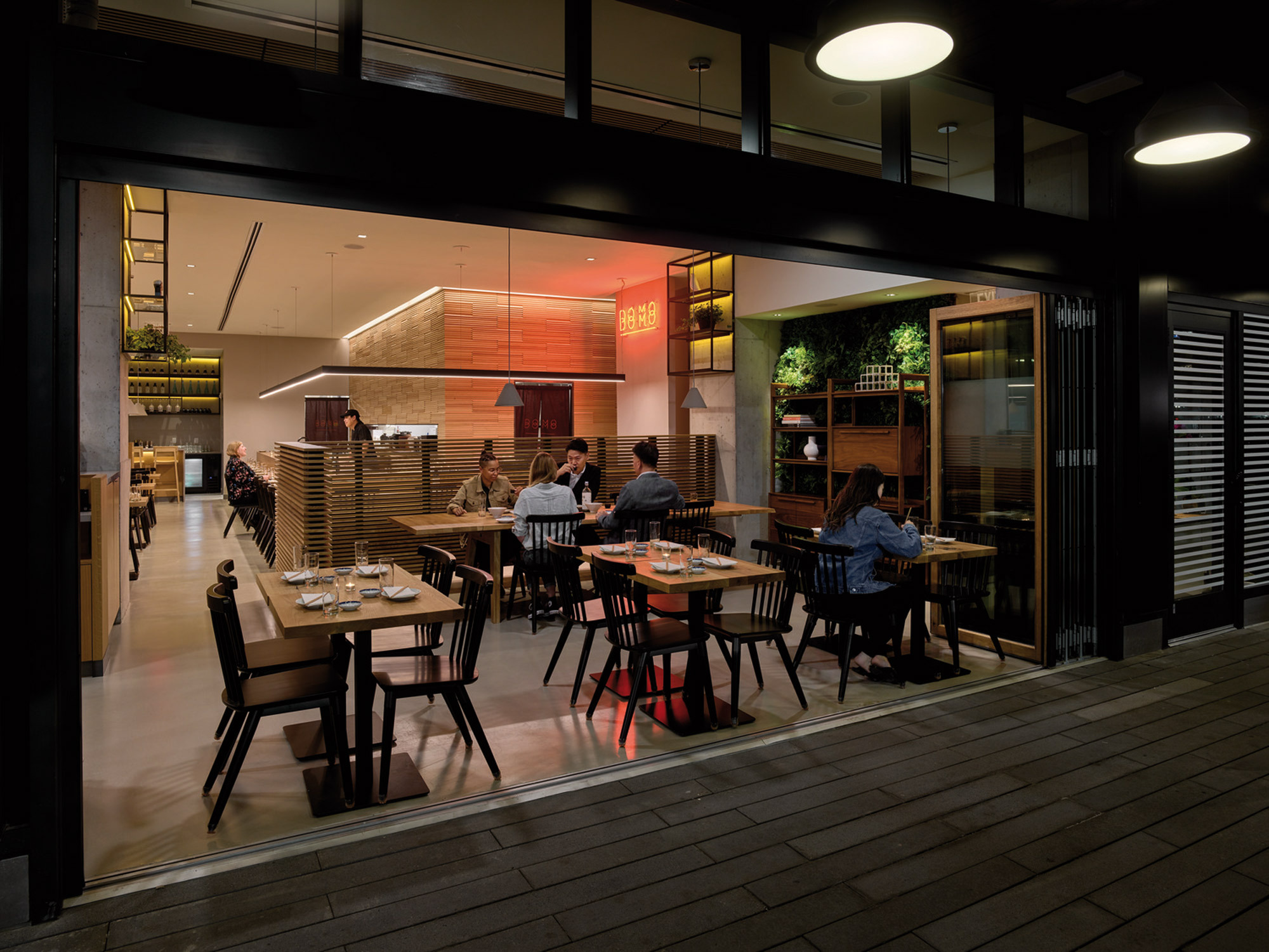 Modern restaurant interior with patrons dining, featuring sleek black framing, warm wood accents, ambient lighting, and a cohesive blend of natural and industrial design elements.