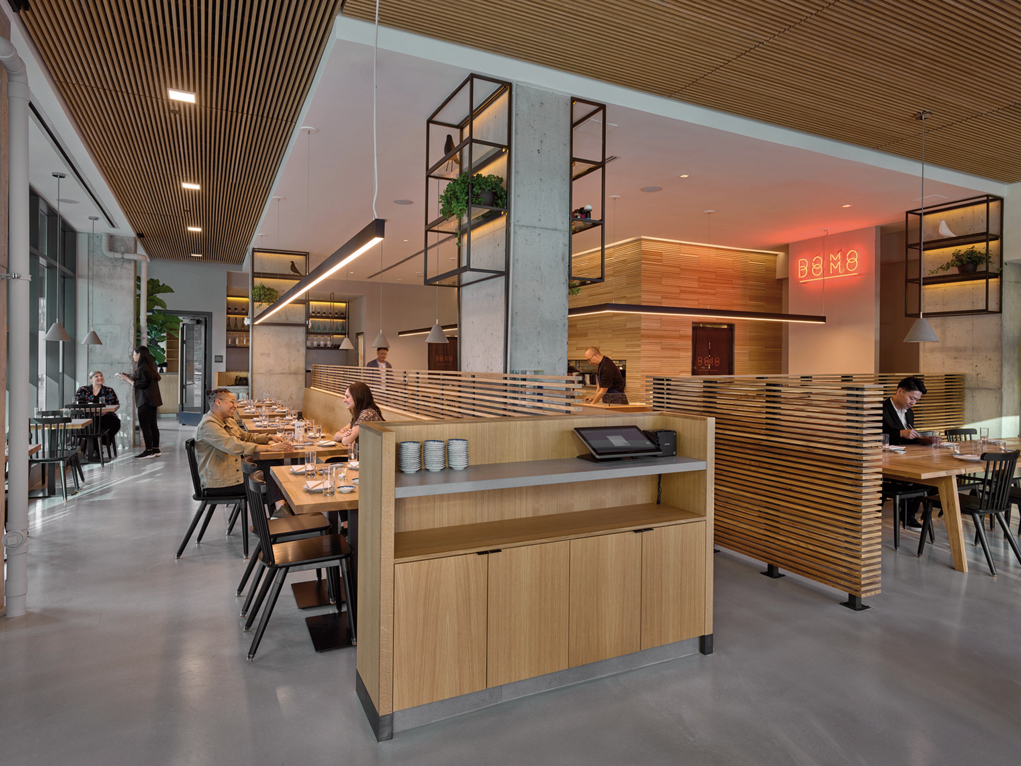 Modern restaurant interior featuring sleek wood paneling, pendant lighting, and exposed concrete columns with greenery. Balanced natural and artificial light complement the warm tones and linear design elements, creating an inviting, contemporary dining atmosphere.