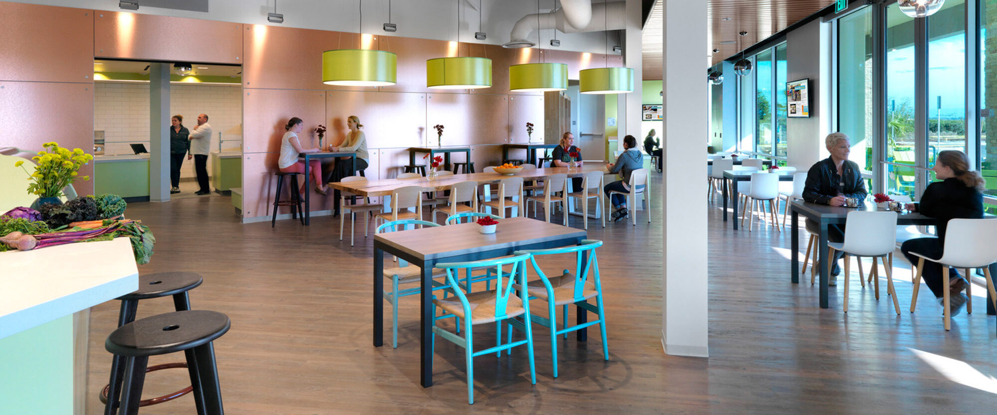Bright and spacious cafeteria featuring sleek white furniture, colorful accents, and wooden cladding. Overhead, pistachio green pendant lights complement the room's modern and welcoming aesthetic. Large windows invite ample natural light, enhancing the open and dynamic dining area.