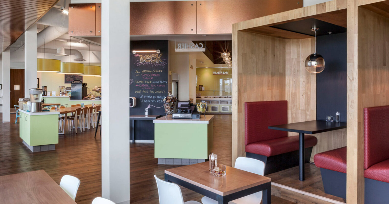Modern café interior showcasing an open-concept layout with a harmonious blend of natural wood tones and vibrant accent colors. Structured wooden beams lead to a cozy dining nook with red upholstered seating. A central serving bar highlights minimalist design with a welcoming chalkboard menu.
