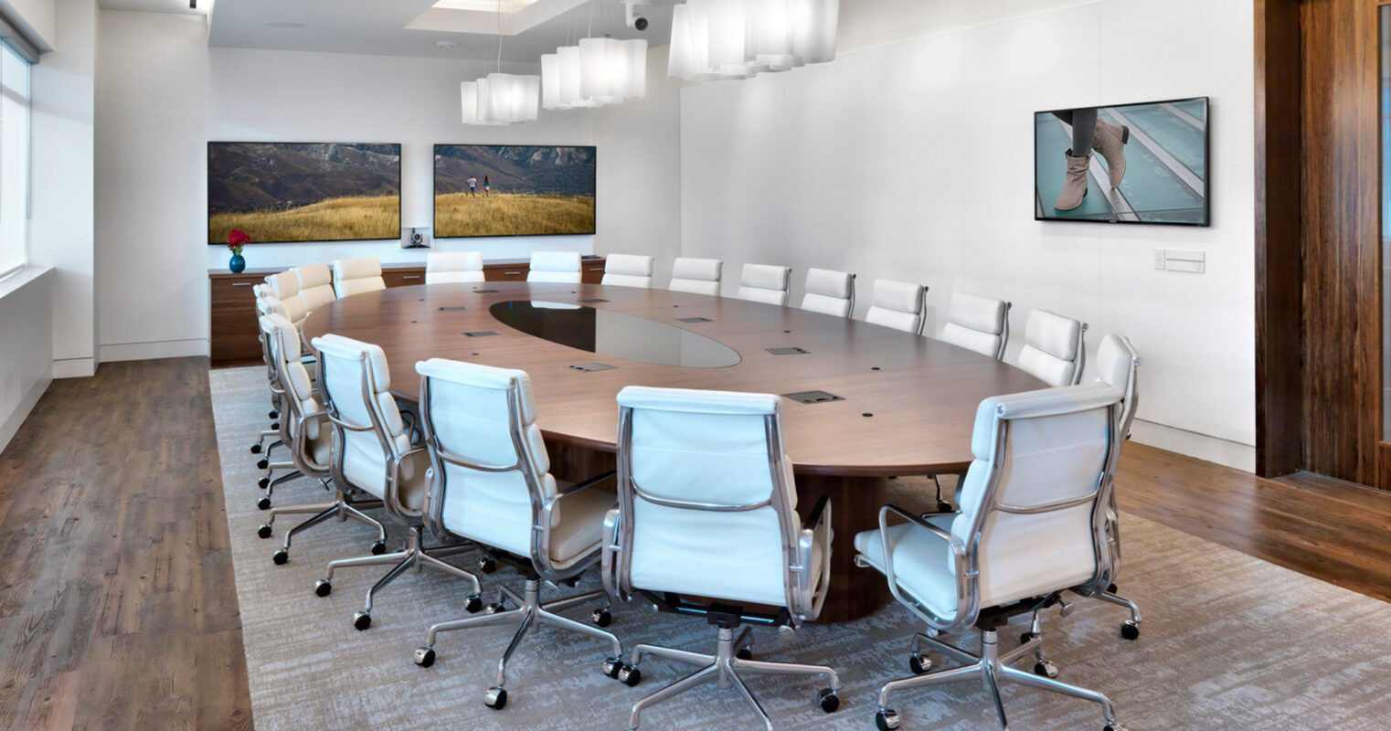 Elegantly appointed conference room featuring a large oval wooden table surrounded by white leather rolling chairs, complemented by a textured neutral carpet. Linear suspension lighting illuminates the space, with serene landscape artworks adorning the walls, all enclosed by warm-toned wooden paneling.