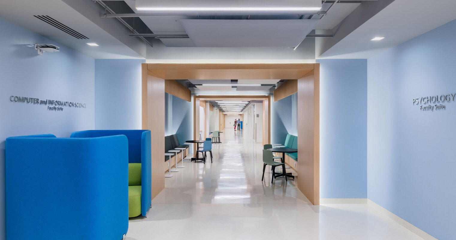 Modern educational facility corridor featuring a vibrant blue and green color scheme, with collaborative seating areas, sleek white flooring, and wall-mounted departmental signage promoting an inviting learning environment.