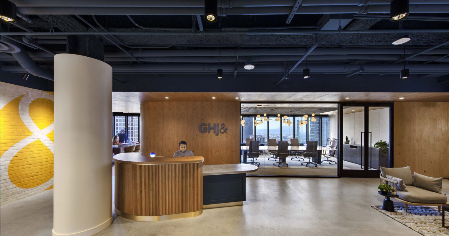 Modern office reception area blending organic and industrial motifs, with a curved wood-paneled desk, sleek pendant lighting, exposed ceiling features, and a monogram logo accent wall. Brickwork adds texture while the adjacent conference room showcases sleek, transparent partitions.