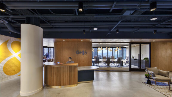 Modern office reception area blending organic and industrial motifs, with a curved wood-paneled desk, sleek pendant lighting, exposed ceiling features, and a monogram logo accent wall. Brickwork adds texture while the adjacent conference room showcases sleek, transparent partitions.