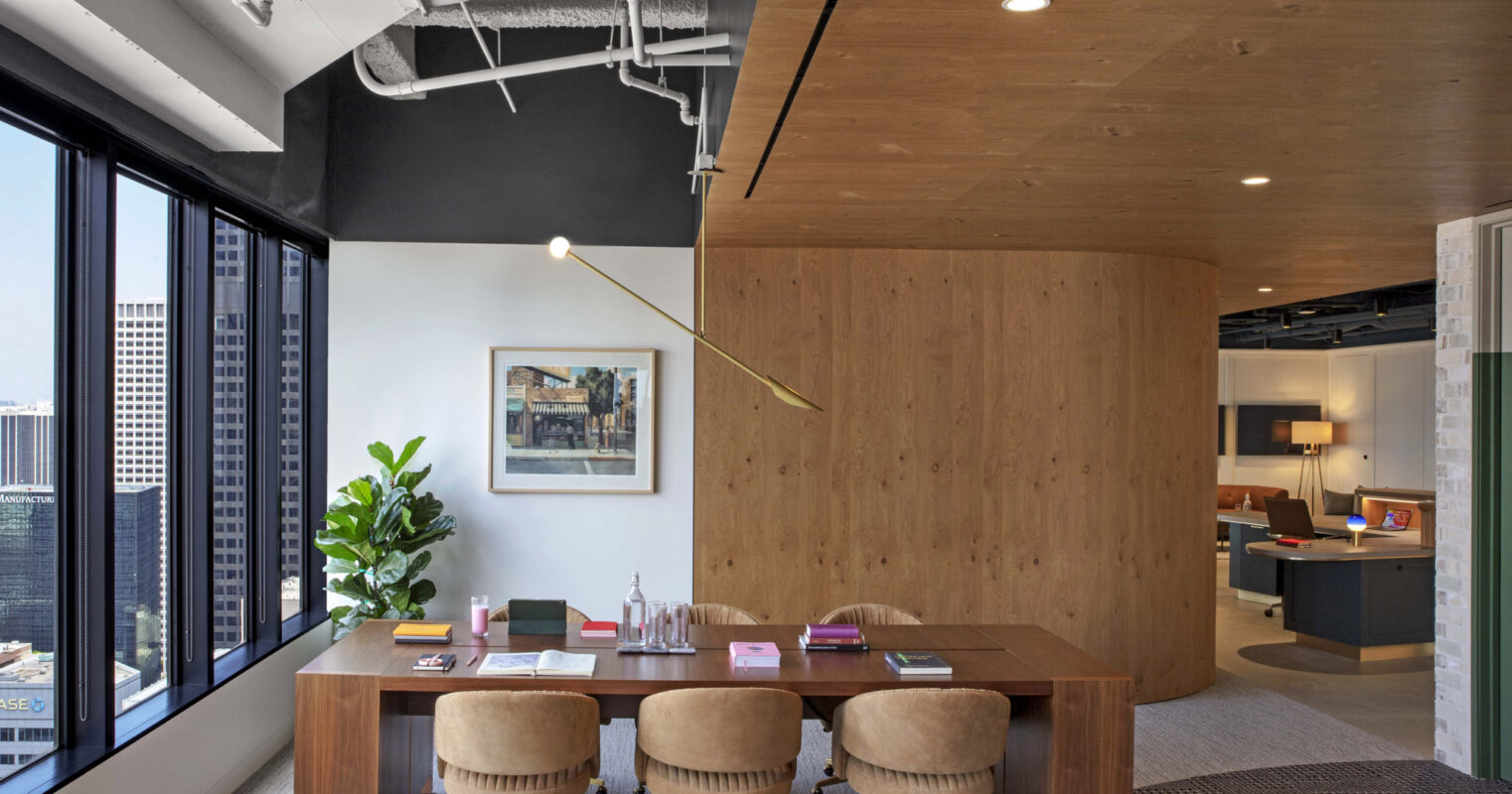 Modern office space with exposed concrete ceiling and ductwork. A warm-toned wooden wall panel contrasts with the industrial look. An oval, wooden table with four plush, swivel armchairs sits atop a textured gray rug, creating a cozy meeting area. Floor-to-ceiling windows offer expansive city views.