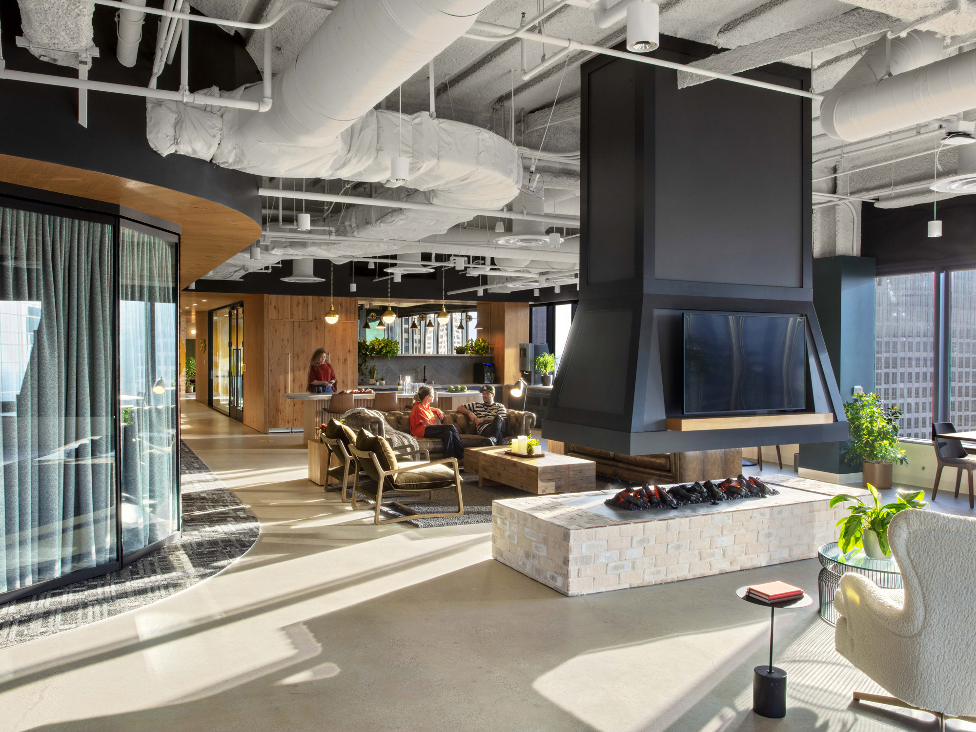 Modern office lounge with exposed ceiling pipes painted white, accented by a central black pendant fireplace. Floor-to-ceiling windows allow natural light, complementing the wood-finished bar area and plants adding a touch of greenery. Plush seating arrangements offer a casual meeting space.