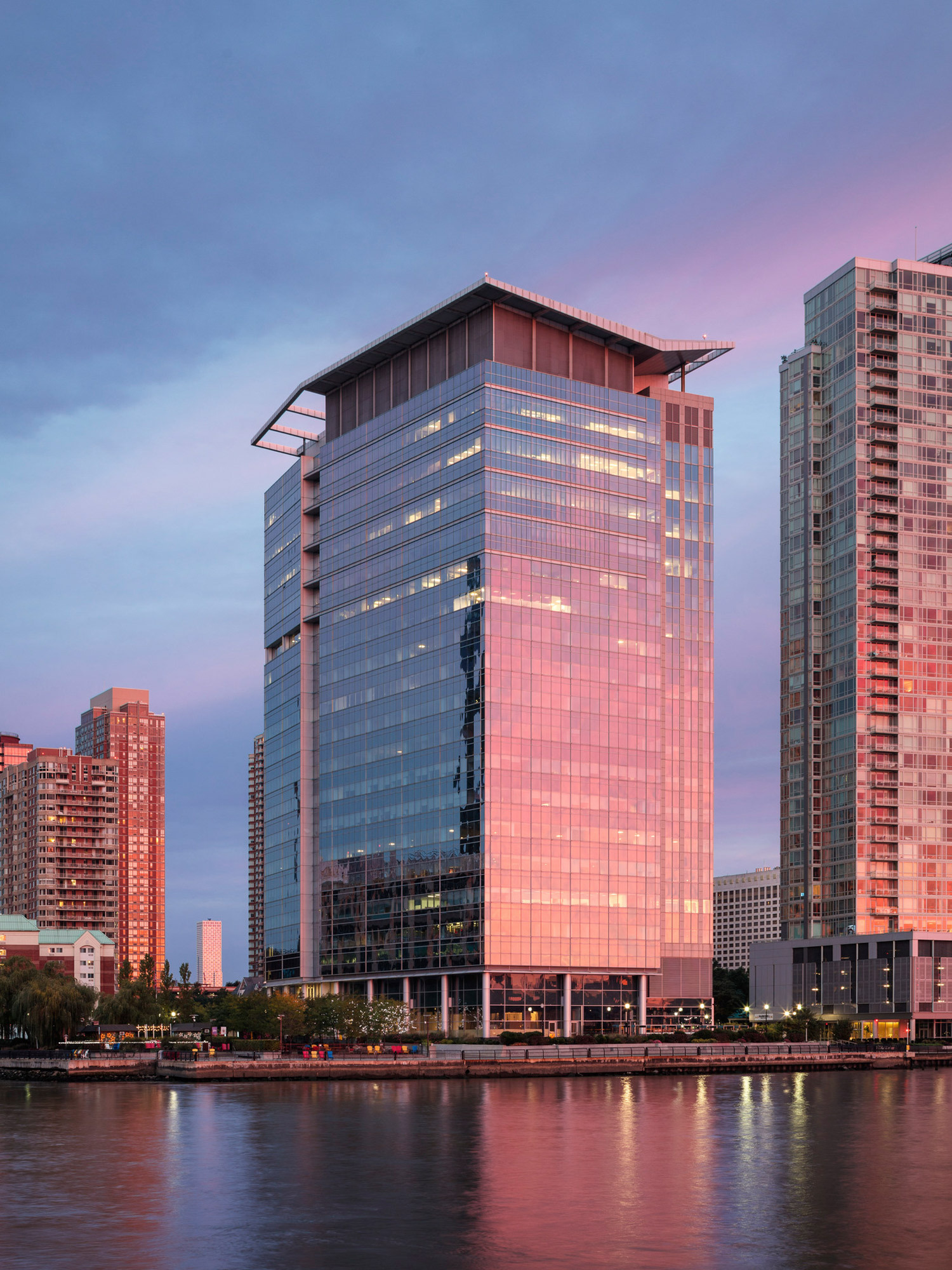 Modern skyscraper with reflective glass facade captures the warm hues of sunset, juxtaposed against a serene river and the soft twilight sky, showcasing contemporary architectural design in an urban waterfront setting.