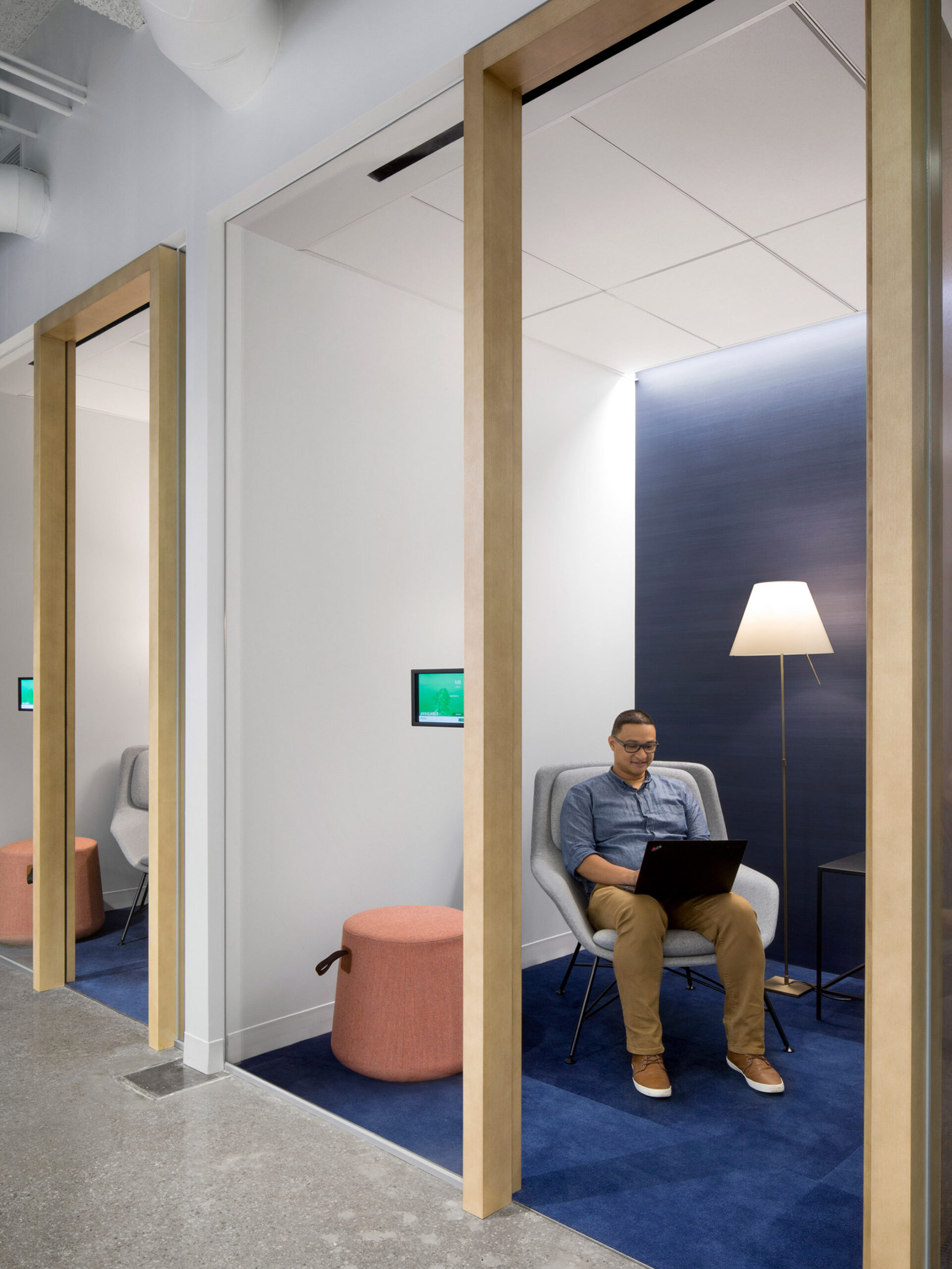 Modern office breakout space featuring glass doors, deep blue acoustic panels, and neutral-toned furniture. A person works on a laptop, seated on a coral ottoman, under a warm floor lamp, highlighting functional yet stylish workplace design.