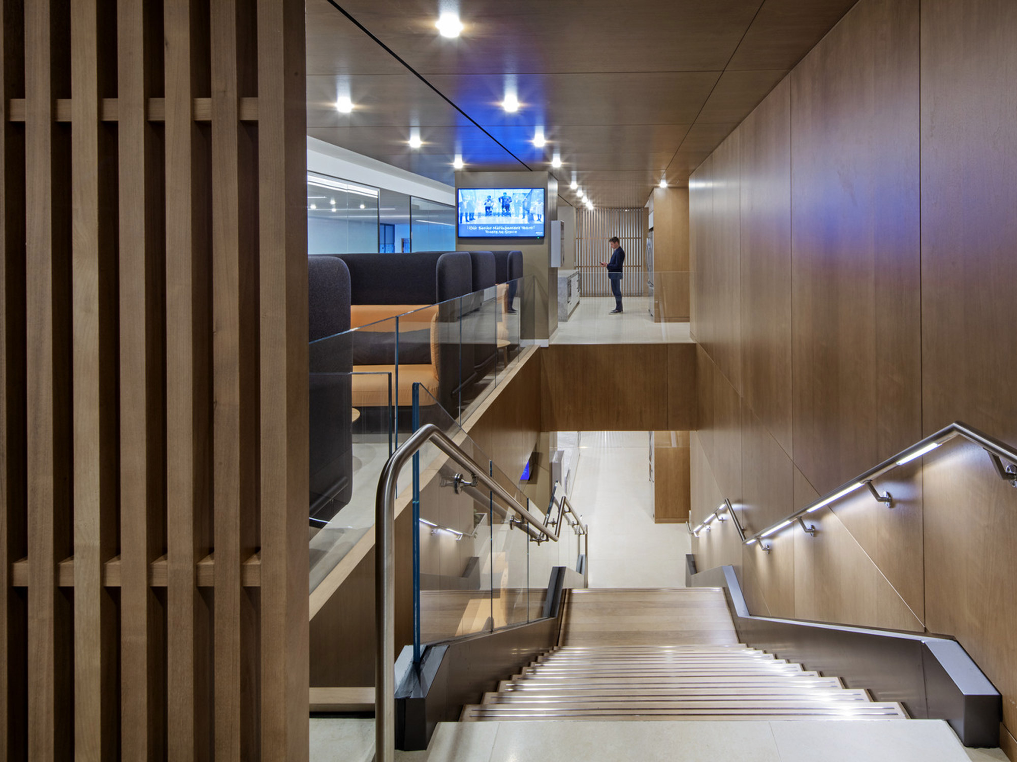Contemporary staircase design integrating wooden panels and glass balustrades, complemented by recessed LED step lighting. The staircase ascends alongside comfortable seating areas with plush blue upholstery, offering a glimpse into a dynamic, well-lit corporate space.