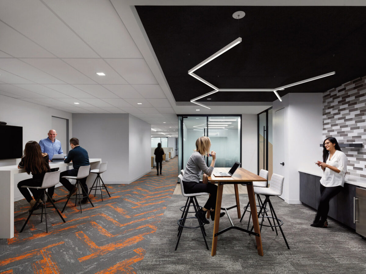 Modern office space with geometric lighting over a collaborative bar-height table, comfortable seating areas, and multitone gray wall tiles. Employees engage in a relaxed meeting environment with vibrant orange carpet accents.