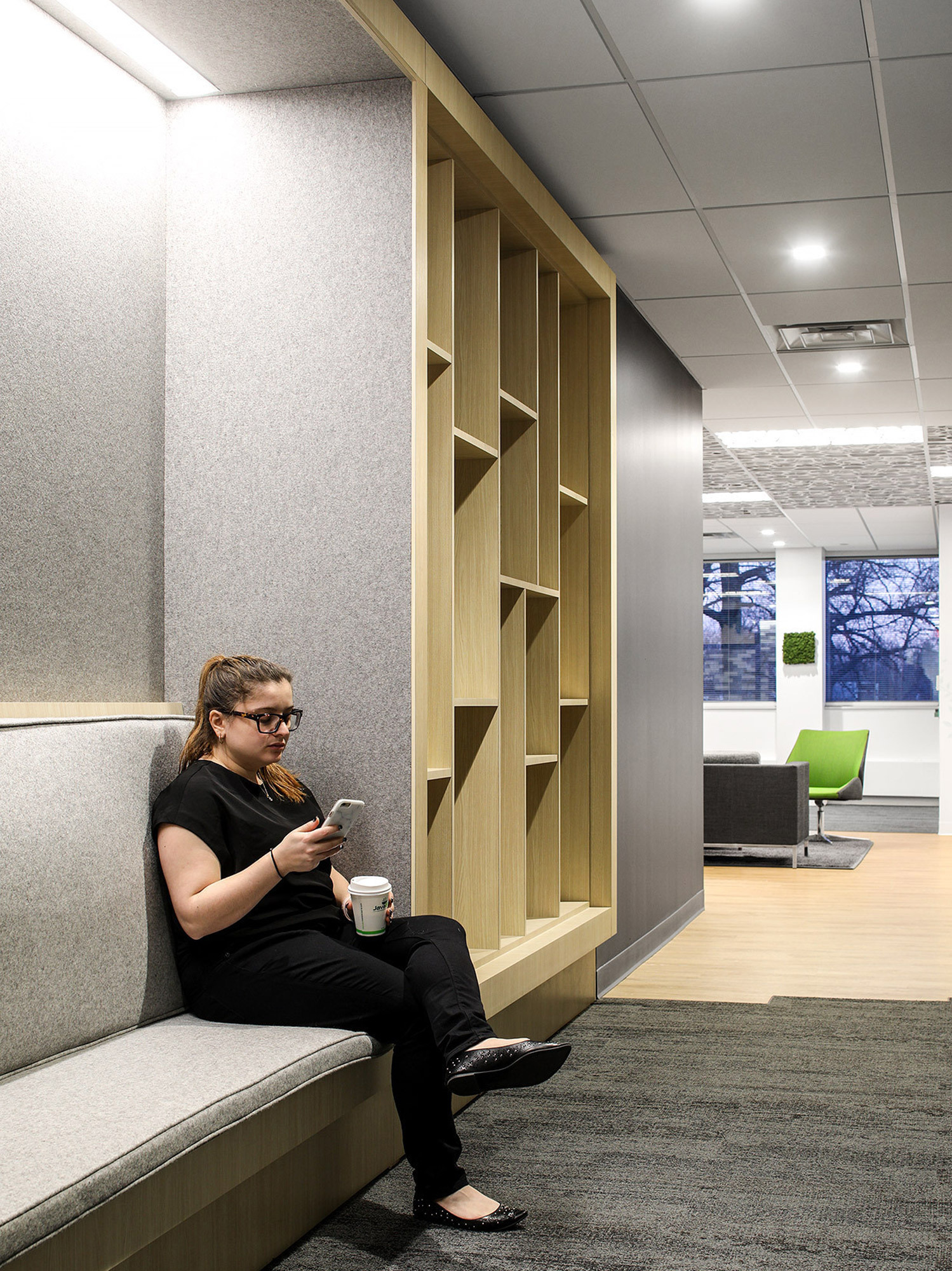 Contemporary office alcove featuring a built-in wooden bookshelf, plush bench seating with gray upholstery, and a person engaged with a smartphone. Subtle tones and textures complement the clean lines, creating a tranquil reading nook with exterior window views.