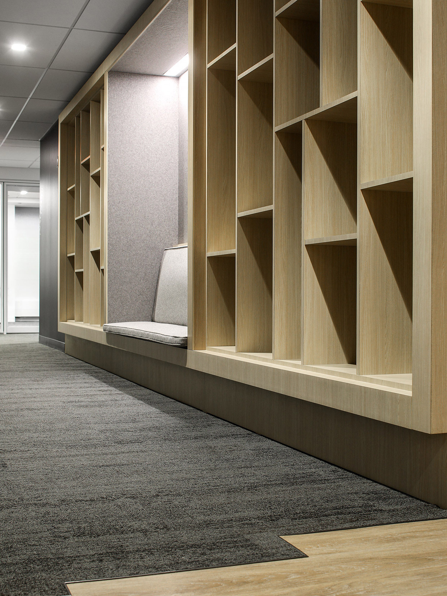 Custom-designed, floor-to-ceiling wooden bookshelves with asymmetrical shelving patterns flank a built-in seating nook, upholstered in light gray. Textured gray carpet flooring transitions to natural wood, highlighting streamlined, modern aesthetics in a corporate office setting.