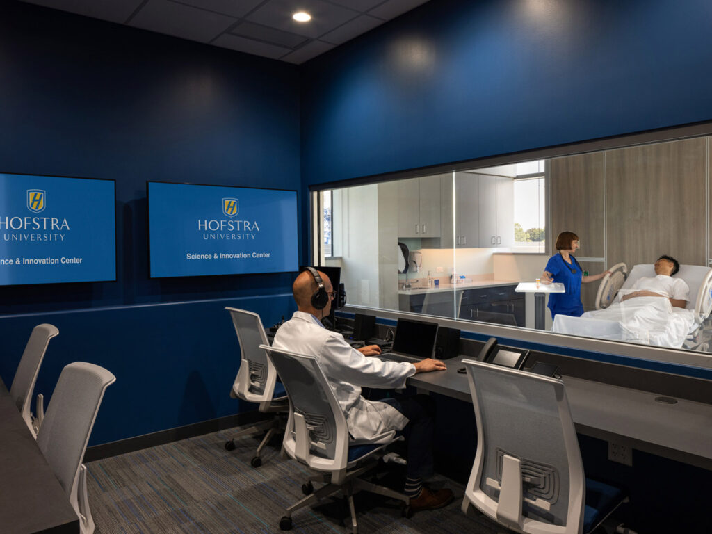 Modern, functional educational facility with observation room featuring ergonomic seating, dual-monitor workstations, glass partition for visibility, and active learning setup with a patient simulation on the adjacent room, showcasing Hofstra University branding on prominently displayed wall signage.