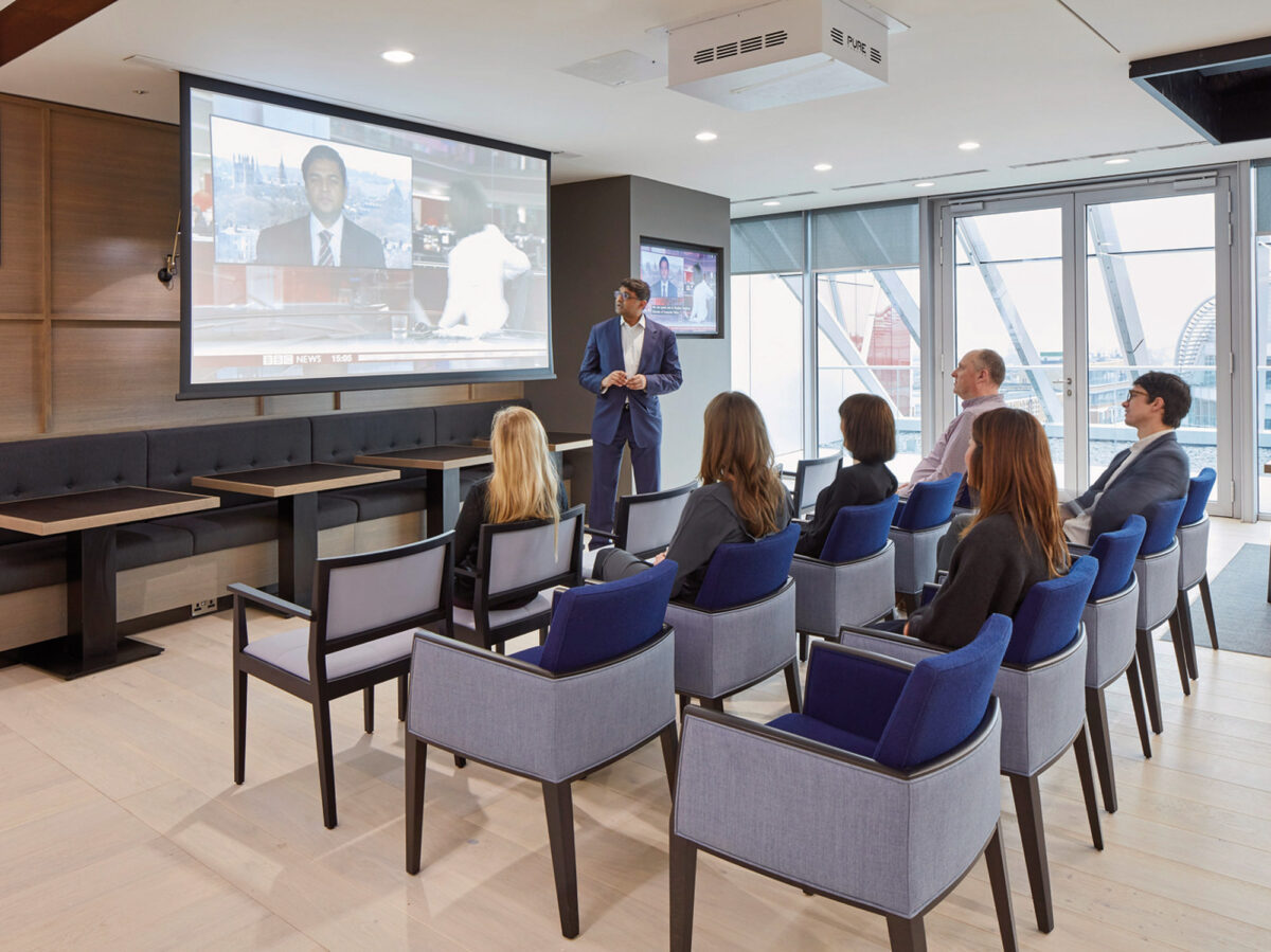 Modern corporate training room with floor-to-ceiling windows offering natural light, ergonomic blue chairs arranged in rows facing a large screen for presentations, and a sleek wooden floor complementing the minimalist aesthetic.