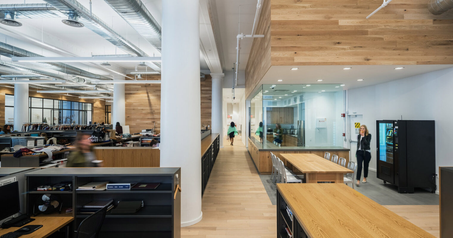 Open-plan office space with natural light featuring exposed ductwork, wooden beams, and communal worktables. Sleek black filing cabinets contrast with light wood floors, enhancing the modern industrial aesthetic.