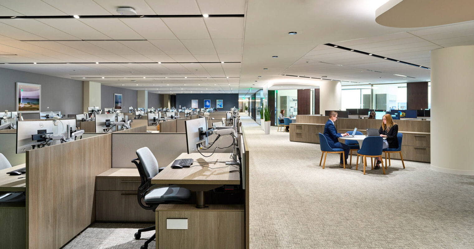 Open-plan office space featuring neutral-toned modular workstations with ergonomic chairs. Overhead, acoustic ceiling panels pair with curved lighting elements. In the foreground, a collaborative area with a wooden table and upholstered blue chairs provides a contrast to the gray carpeted floors.