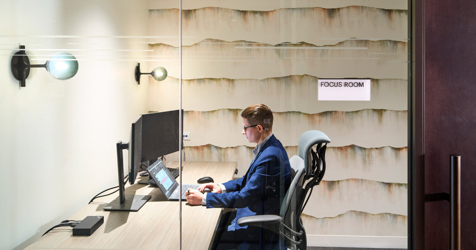 Professional focus room interior featuring a textured, wave-like wall panel in a creamy palette, modern wood-grain desk, ergonomic black chair, and glass partition, complemented by ambient lighting fixtures.