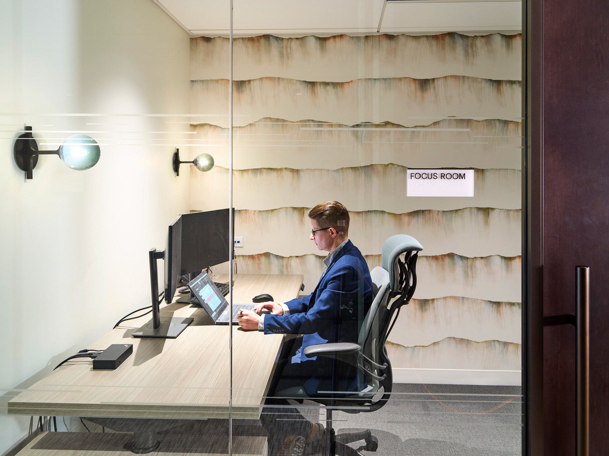 Professional focus room interior featuring a textured, wave-like wall panel in a creamy palette, modern wood-grain desk, ergonomic black chair, and glass partition, complemented by ambient lighting fixtures.