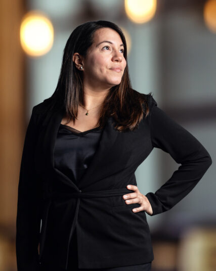 The image depicts a woman with long dark hair and a contemplative expression, dressed in a black blazer and top, with her hand on her hip. The background features warm, circular bokeh lights, which contribute to a sophisticated atmosphere likely within an office setting. The image depicts a woman with long dark hair and a contemplative expression, dressed in a black blazer and top, with her hand on her hip. The background features warm, circular bokeh lights, which contribute to a sophisticated atmosphere likely within an office setting. The image depicts a woman with long dark hair and a contemplative expression, dressed in a black blazer and top, with her hand on her hip. The background features warm, circular bokeh lights, which contribute to a sophisticated atmosphere likely within an office setting.