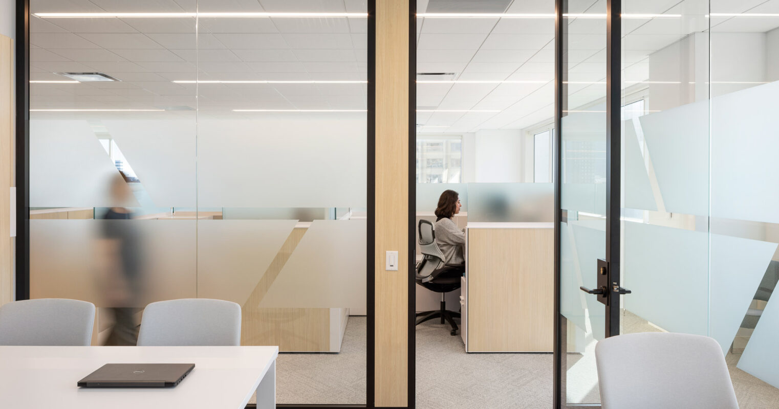Modern office meeting room with frosted glass partitions, light wood finishes, and ergonomic chairs, alongside a view into an individual focused workspace, symbolizing privacy and collaboration in a corporate setting.