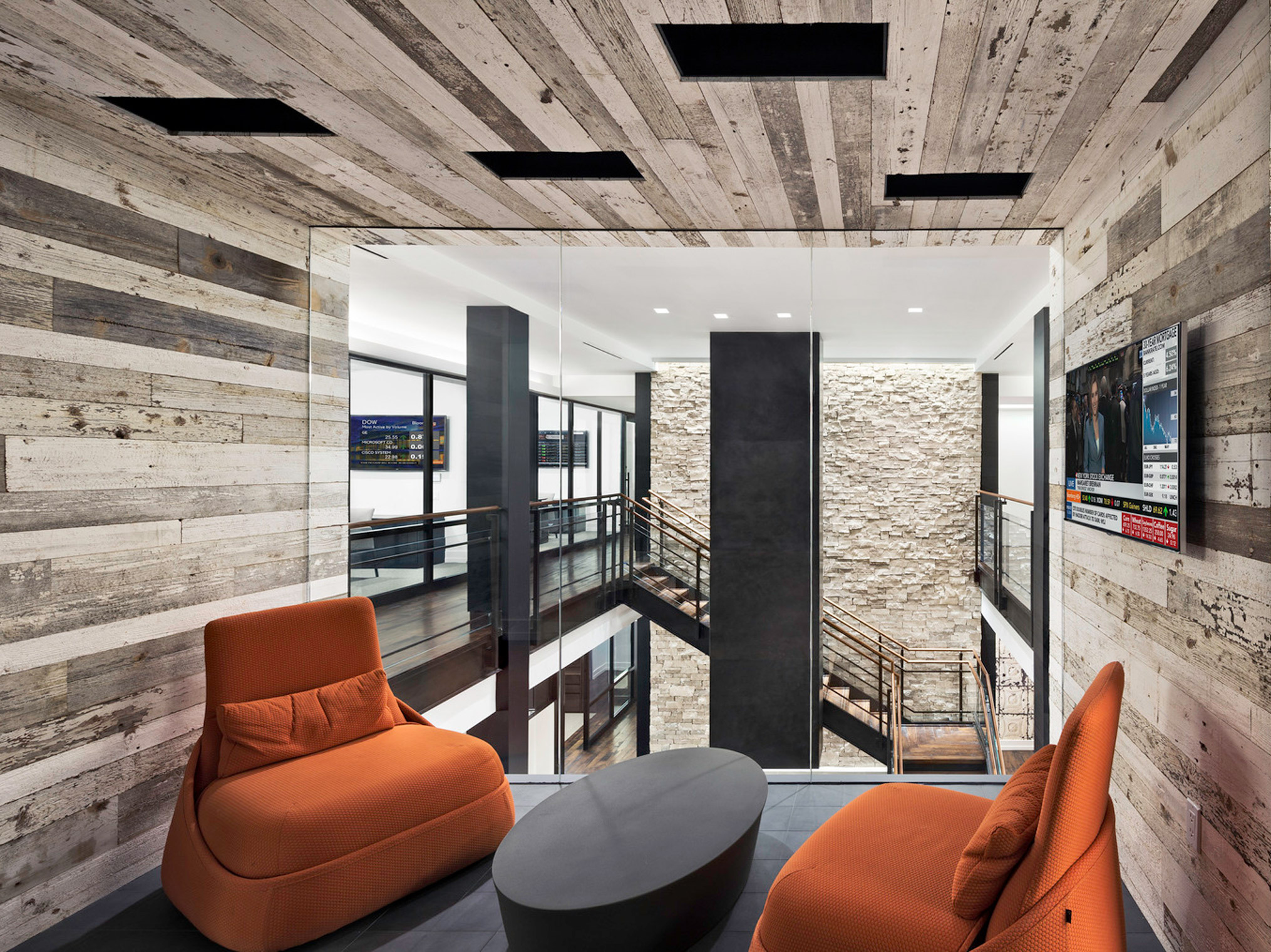 Modern open-plan interior featuring reclaimed wood-clad walls and ceilings juxtaposed with sleek black beams and railings. Burnt orange armchairs and a slate gray ottoman create a warm, inviting focal point before expansive glass panes overlooking a staircase and stone column.