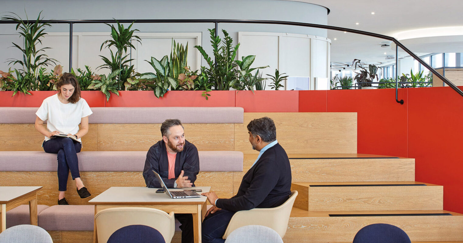 Open-plan office with layered seating arrangement, featuring pink and neutral-hued upholstery, wooden bleachers, and vibrant red partitions. Greenery accents the space with a sense of biophilic design, fostering a collaborative and relaxed working environment.
