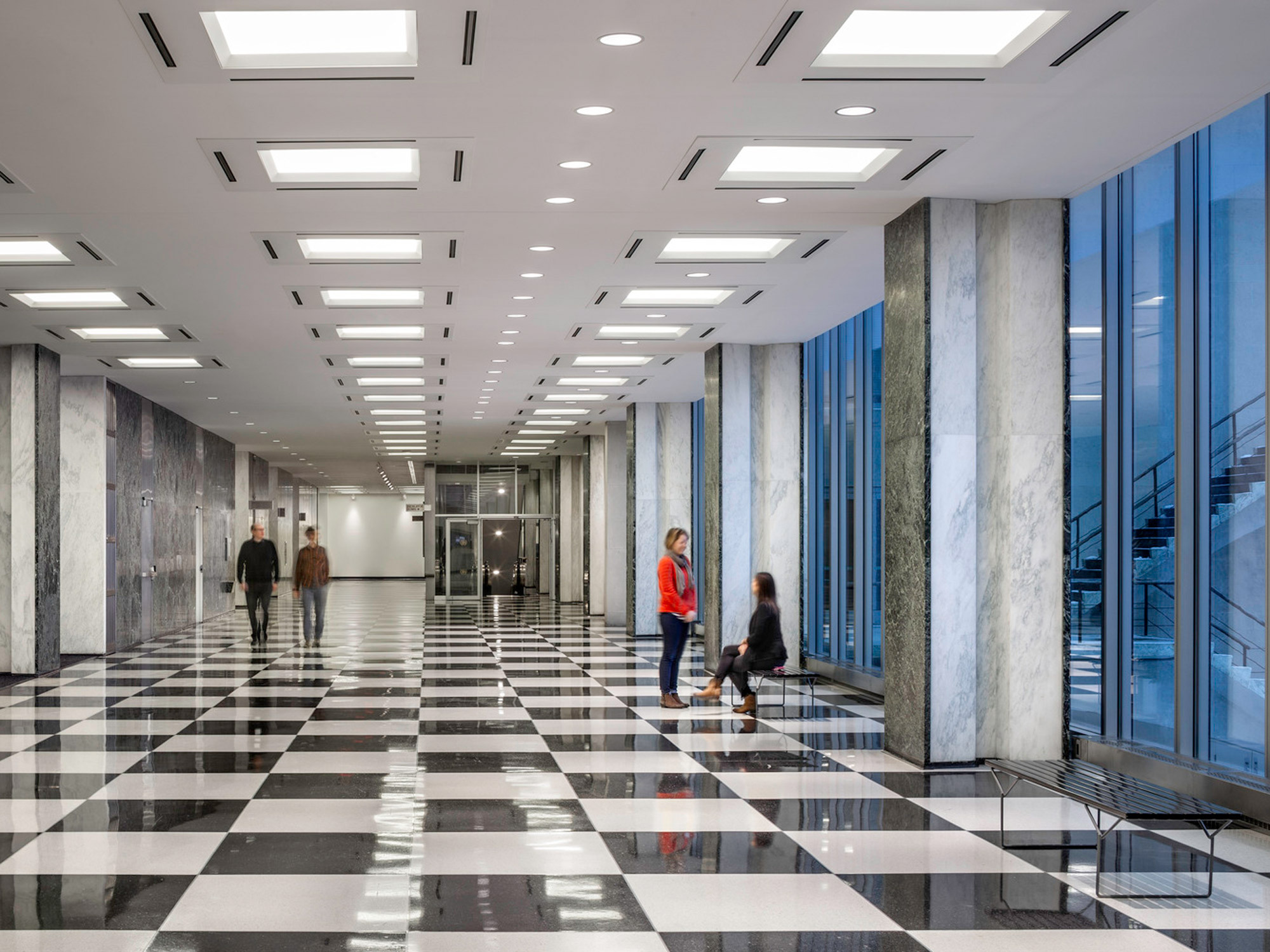 Modern commercial lobby with monochromatic checkered flooring, sleek marble columns, ample ceiling-mounted lighting fixtures, and full-height glass windows emitting natural light. People are walking and conversing, adding human scale to the space.
