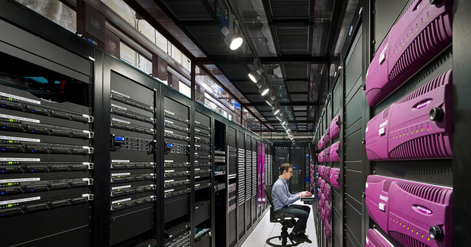 Modern data center interior with rows of purple server racks, leading lines creating depth, and cool lighting highlighting technological infrastructure. A professional sits attentively at a workstation, monitoring system performance and data integrity in a space optimized for efficiency and security.
