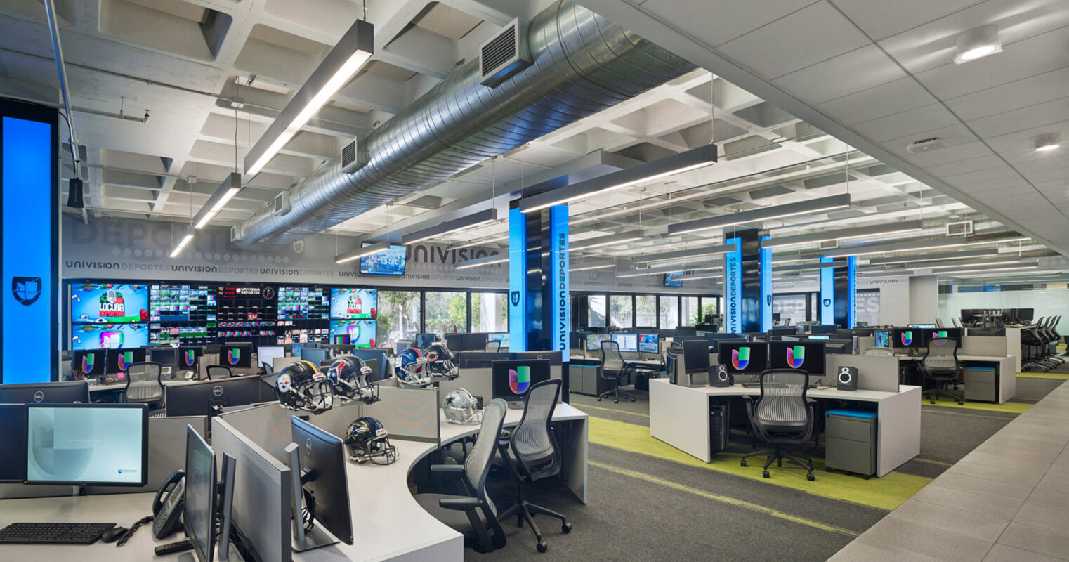 Modern office space featuring an open-floor plan with clusters of ergonomic workstations under exposed ductwork, infused with natural light from expansive windows, and accented by vibrant blue structural columns. Dynamic digital screens add a technological edge to the environment.