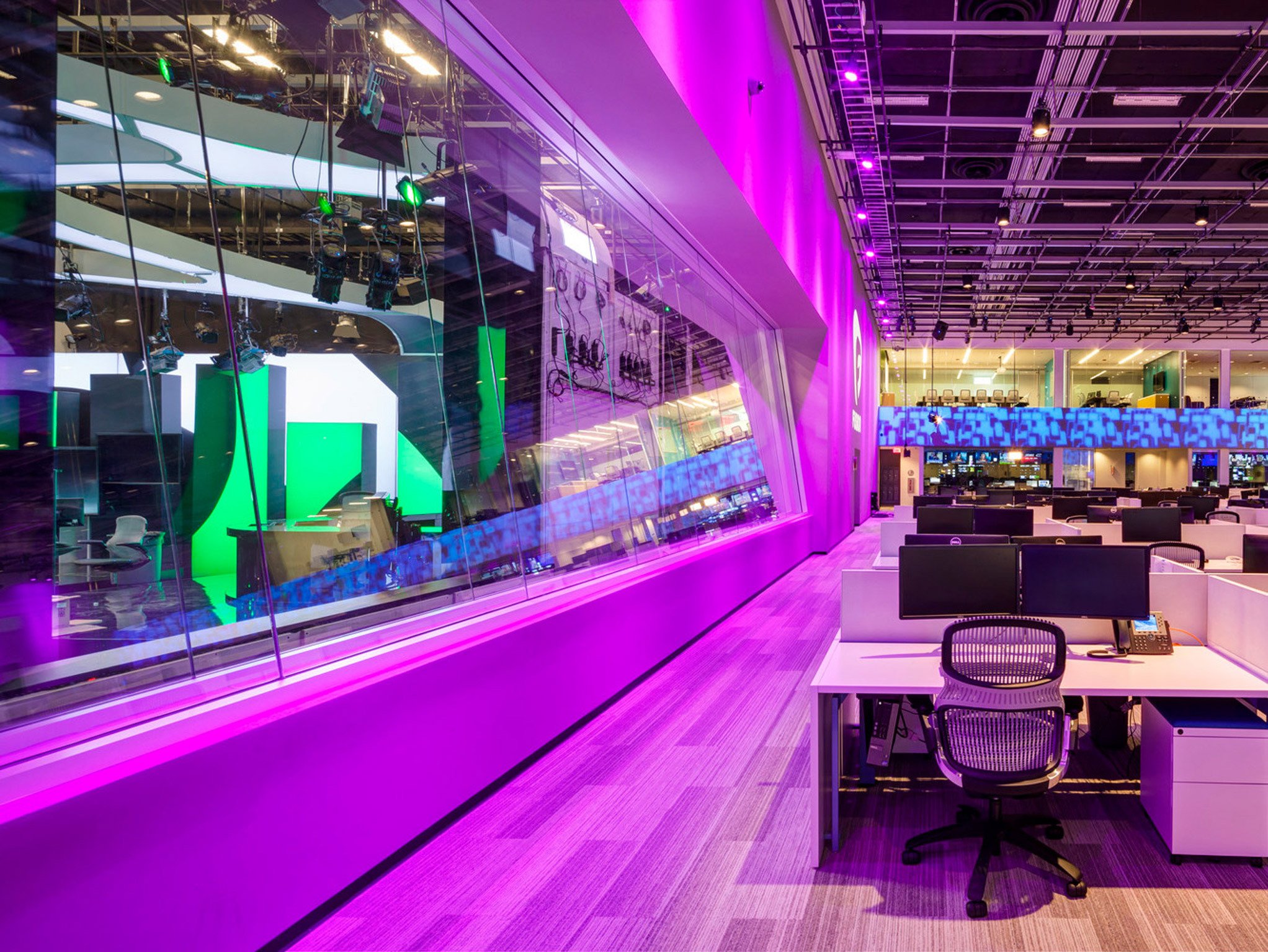 Modern open-plan office space with vibrant pink accent lighting along the windowed wall. It features sleek white desks, ergonomic black chairs, and modular overhead lighting grid. The environment is enhanced by a backdrop of dynamic multi-screen video installation.