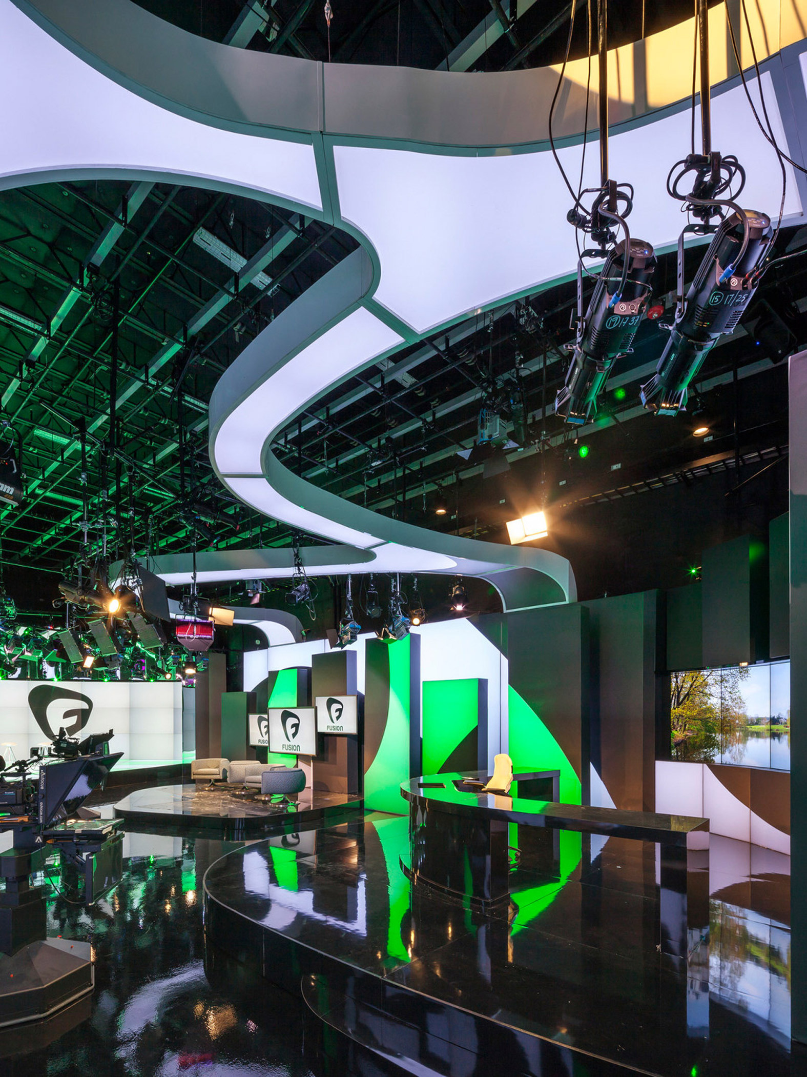 Sleek, modern broadcasting studio featuring a dynamic curved white overhead structure, integrated LED lighting, and green accent hues. Multiple monitors and contemporary furniture populate the space, with advanced camera rigs suspended from the ceiling for versatile filming angles.