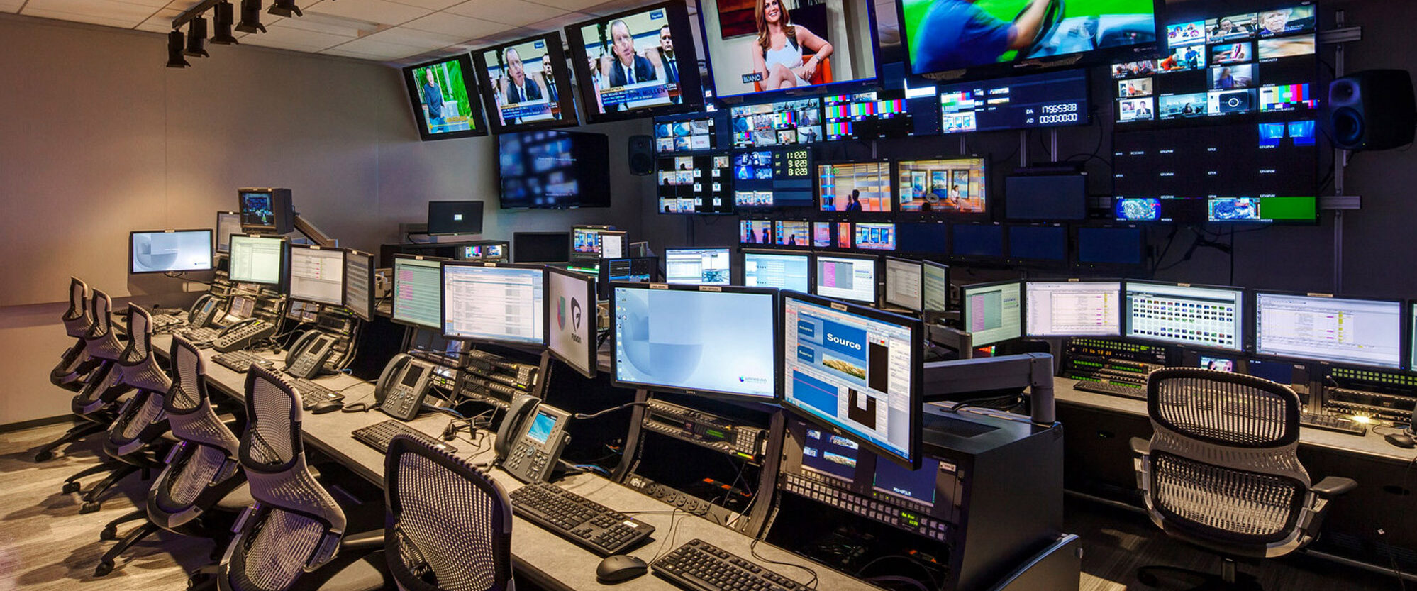 Modern broadcast control room lined with multi-screen display panels. Ergonomic chairs and sophisticated audio-visual equipment adorn the space, reflecting a high-tech, efficient workplace designed for media monitoring and production tasks.