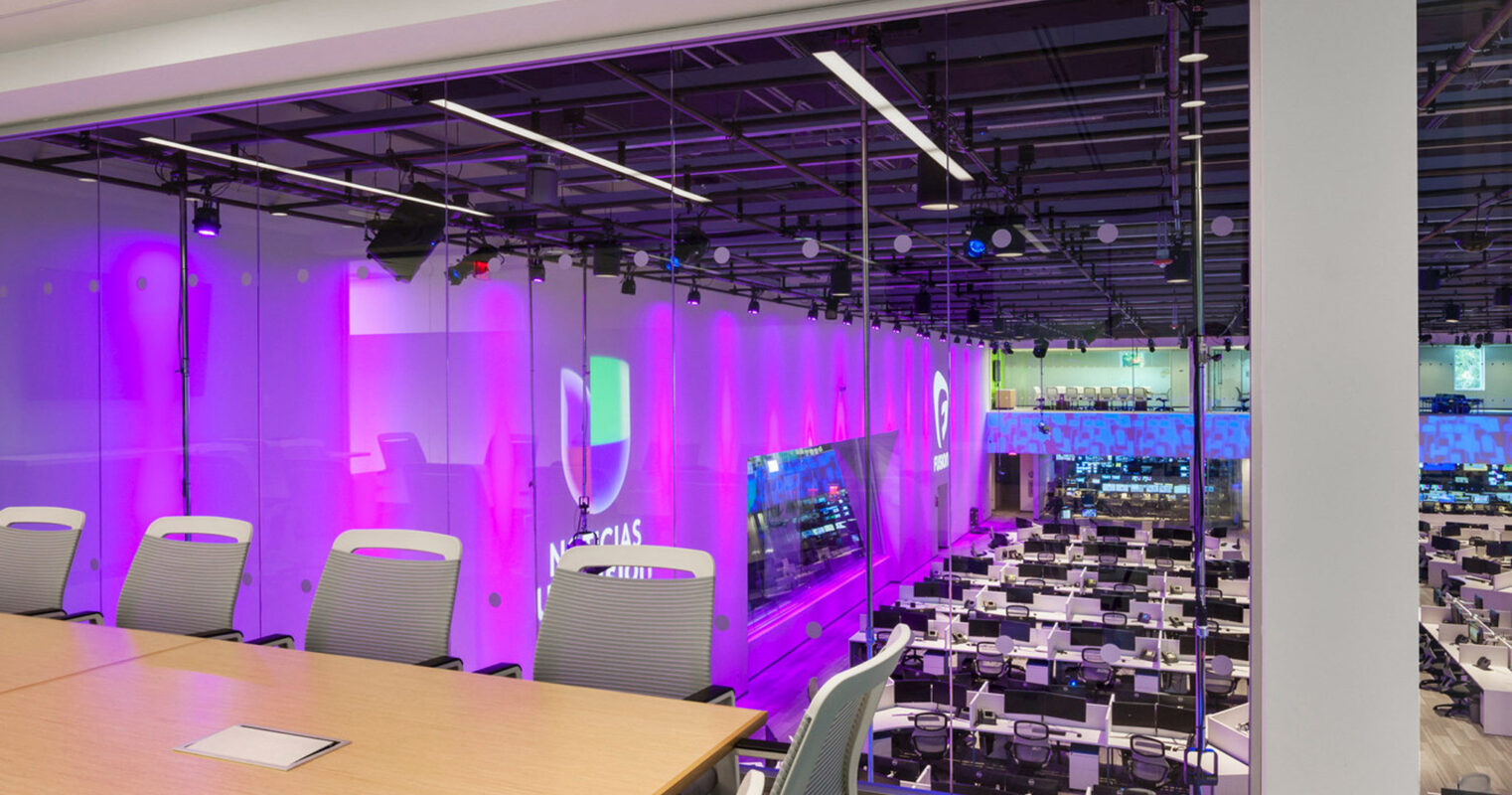 Modern office conference room with a clear glass wall overlooking a bustling open-plan workspace featuring ergonomic chairs and neatly arranged workstations. Vibrant purple LED lighting accents the adjacent media broadcasting room, showcasing contemporary corporate design with a focus on functionality and brand identity.